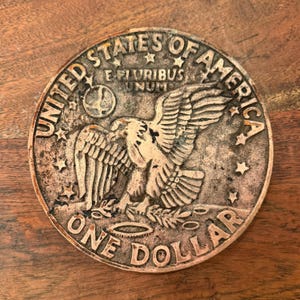 May include: A silver-coloured metal coin with the words "United States of America" and "One Dollar" around the edge. The coin features an eagle with outstretched wings and the words "E Pluribus Unum" above it.