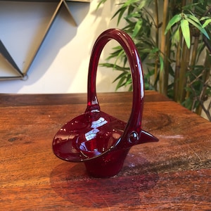 May include: A small, red glass basket with a handle. The basket is made of clear glass with a red tint. It is sitting on a wooden surface.