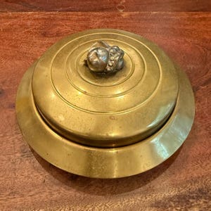 May include: A round, antique brass container with a lid. The lid features concentric circles and a small, ornate, silver-colored knob. The container sits on a wooden surface, showcasing its aged patina and vintage charm.