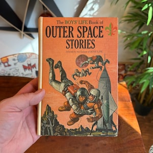 Puede incluir: Portada de libro vintage titulada "The BOYS' LIFE Book of OUTER SPACE STORIES". La portada es naranja y presenta ilustraciones de astronautas, un cohete y planetas. El libro está sostenido por una persona.