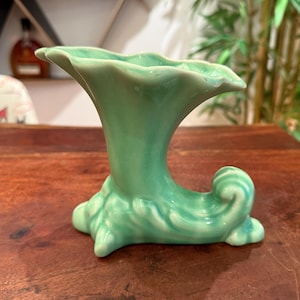 May include: A light green ceramic cornucopia vase featuring a glossy finish and a flared, scalloped rim. The base of the vessel is decorated with molded, swirling leaf-like patterns that curve upward into a horn-shaped opening.