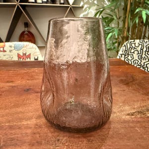 Può includere: Un vaso di vetro trasparente con una superficie strutturata e una base ampia. Il vaso ha una sottile tonalità rosata e una parte superiore leggermente svasata. È esposto su un tavolo di legno, con uno sfondo sfocato.