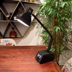 Op de afbeelding: Een zwarte bureaulamp met een verstelbare arm en een rechthoekige voet. De lamp heeft een klassiek ontwerp met een lampenkap die het licht naar beneden richt. Hij staat op een houten tafel.