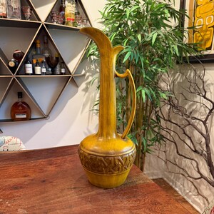 Puede incluir: Una jarra alta de cerámica amarillo mostaza con cuello delgado y asa curva. La base presenta un patrón geométrico en relieve detallado. La jarra se exhibe sobre una superficie de madera, mostrando su diseño vintage.