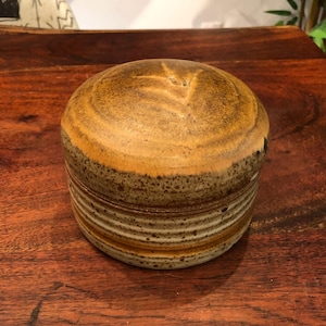 Może przedstawiać: Okrągły pojemnik ceramiczny z pokrywką. Pojemnik jest brązowy i biały z pstrokatą glazurą. Pokrywka ma subtelny wzór liścia.