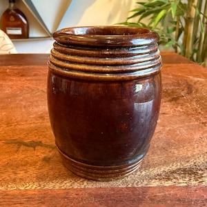 May include: A brown ceramic jar with a cylindrical shape and a series of horizontal ridges around the top. The jar has a glossy finish and appears to be handmade. The jar is sitting on a wooden surface.