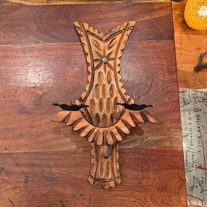 May include: A wall-mounted wooden candle holder featuring intricate hand-carved patterns, including a central star motif and scalloped edges. The piece has two black metal candle cups extending from the sides and a light brown wood finish.