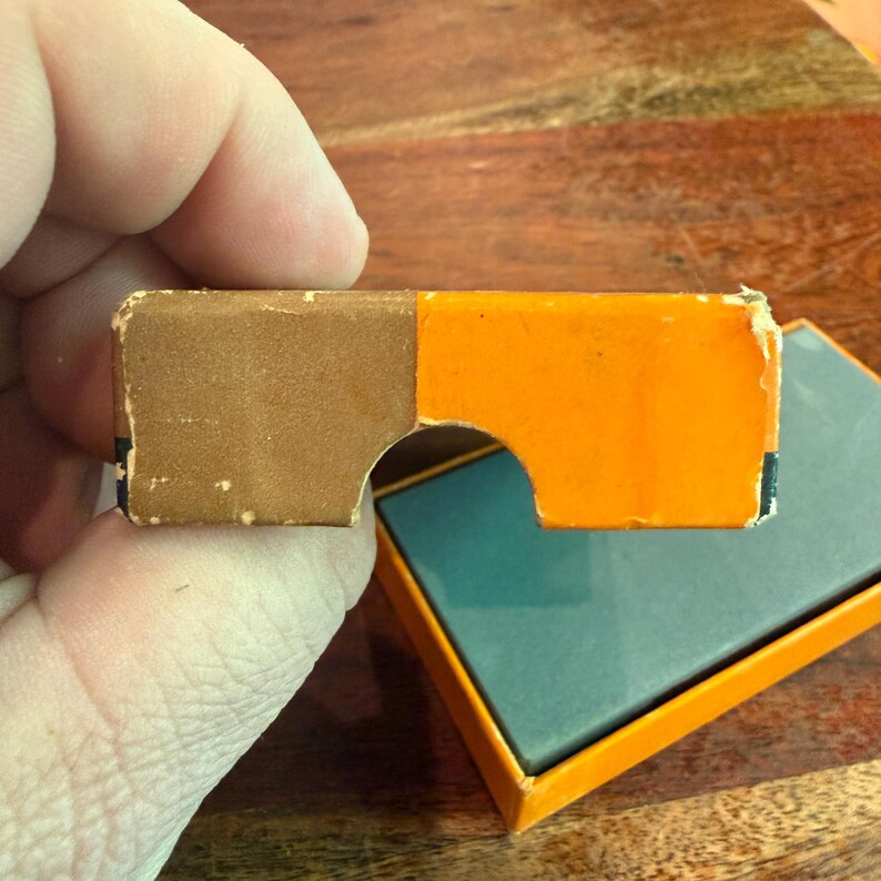 May include: A vintage cardboard viewer with a brown and orange color scheme. The viewer has a cutout section and is partially open, revealing a teal interior. The item is likely a retro toy or collectible.