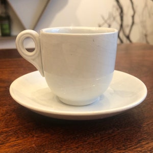 May include: A white ceramic coffee cup and saucer set. The cup has a rounded shape and a small handle. The saucer is a simple, round shape.