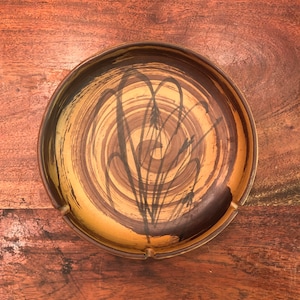 May include: A brown ceramic ashtray with a swirl pattern and black brushstrokes. The design resembles stylized grass or reeds.