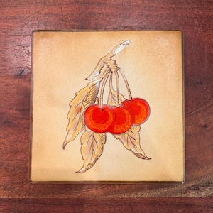 May include: A square ceramic tile featuring a central illustration of three bright red cherries hanging from a single stem. The cherries are surrounded by stylized, light tan leaves with dark brown outlines, set against a smooth, beige background.