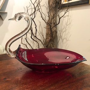 May include: A clear glass swan-shaped serving dish with a deep red bowl. The swan's neck is curved and the bowl is heart-shaped.