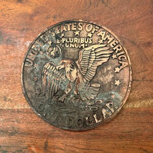 May include: A copper-coloured one dollar coin with the words "United States of America" and "One Dollar" around the edge. The coin features an eagle with its wings spread, and the words "E Pluribus Unum" are inscribed above the eagle.