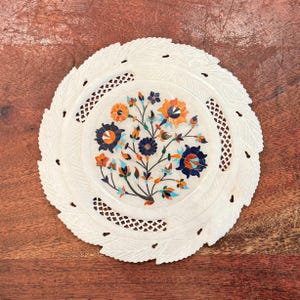 May include: A white, round decorative plate with a floral inlay design. The plate features a detailed pattern of flowers in shades of orange, blue, and green. The outer edge has a leaf-like design. The plate is set on a wooden surface.