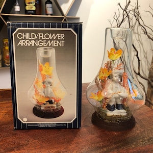 Diorama de porcelana kitsch de niño y flor artificial de c. 1983 en campana de cristal sobre soporte con caja de Action Industries - Decoración kitsch de los años 80