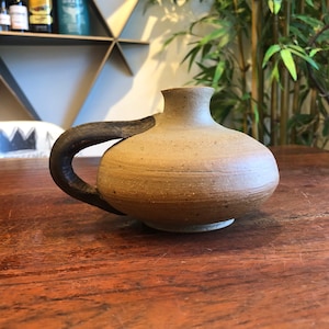 Puede incluir: Un pequeño jarrón redondo de cerámica beige con un asa marrón oscuro. El jarrón tiene una textura moteada y una superficie ligeramente irregular.