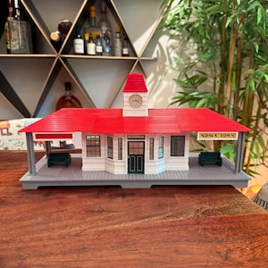 May include: A model train station building with a bright red roof, white walls, and a central clock tower. The structure features a grey platform, dark green benches, and a sign that reads NOMA TOWN. The building has multiple windows and a door.
