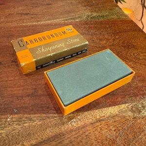 May include: A vintage Carborundum brand sharpening stone in its original box. The box is brown and orange with the words "Carborundum Sharpening Stone" printed on it. The sharpening stone is gray and rectangular, resting in an orange tray.