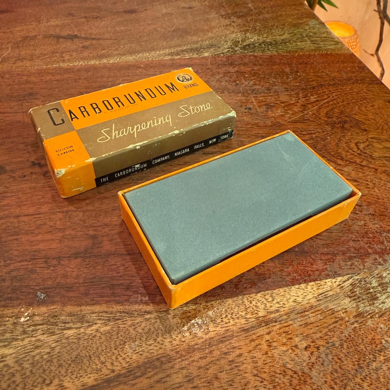 May include: A vintage Carborundum brand sharpening stone in its original box. The box is brown and orange with the words "Carborundum Sharpening Stone" printed on it. The sharpening stone is gray and rectangular, resting in an orange tray.