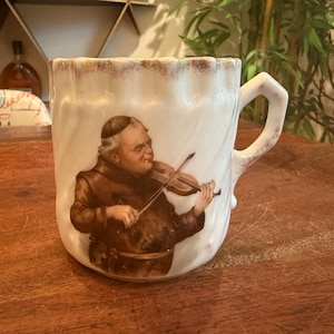 Op de afbeelding: Een witte keramische mok met een gedetailleerde schildering van een man die viool speelt. De mok heeft een decoratieve rand en een gebogen handvat. De man op de schildering draagt een bruine jurk en een zwarte hoed.