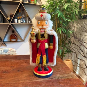 May include: A decorative wooden nutcracker in the shape of a king, standing on a round base. The king wears a gold and red outfit with a white fur-trimmed cape, a crown, and holds a scepter. The nutcracker is approximately 12 inches tall.