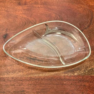 Puede incluir: Plato de servir de vidrio transparente en forma de hoja con un acento de metal plateado. El plato está dividido en dos secciones por un divisor de vidrio curvo. El plato está sobre una superficie de madera.