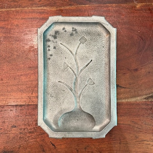 Könnte beinhalten: Ein rechteckiges Metalltablett mit einem stilisierten Blumendesign. Das Tablett hat eine strukturierte Oberfläche und einen erhöhten Rand. Das Blumendesign zeigt eine Vase mit einem Stiel und drei blütenartigen Formen. Das Tablett ist silberfarben.
