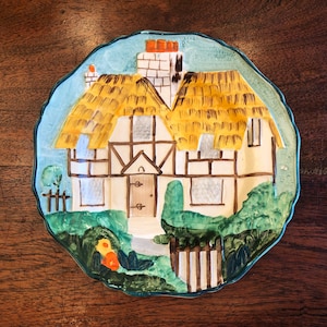 Puede incluir: Un plato de cerámica vintage con un diseño pintado a mano de una cabaña con techo de paja, exterior blanco y molduras marrones. La cabaña está rodeada de vegetación verde y una valla de madera.