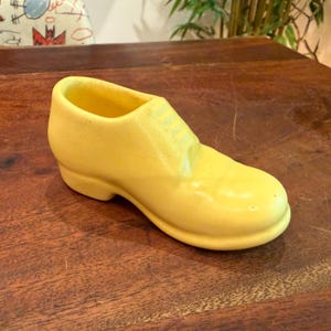 May include: A yellow ceramic shoe-shaped planter. The planter is a light yellow color and has a classic shoe design with a rounded toe and a small heel. The opening is at the top of the shoe.