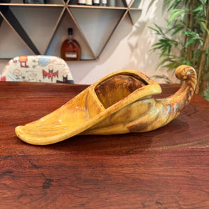 May include: A ceramic cornucopia-shaped vessel featuring a glossy mustard yellow glaze with dark brown streaking. The object has a wide, flat opening that tapers into a curved, pointed tail, showcasing a smooth, reflective surface throughout.