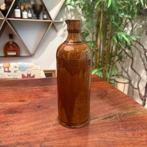 Könnte beinhalten: Eine braune antike Flasche mit schmalem Hals und zylindrischem Körper. Die Flasche hat horizontale geätzte Linien in der Nähe der Oberseite und besteht aus glasierter Keramik. Die Flasche wird auf einer Holzoberfläche ausgestellt.