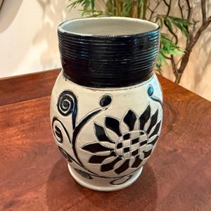 May include: A ceramic vase featuring a cream-colored body with a dark blue, ribbed neck. The exterior is decorated with a hand-carved, dark blue floral pattern consisting of a large central flower and swirling vine motifs on a smooth surface.