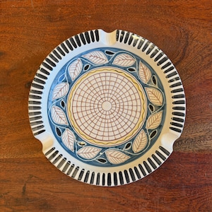 Puede incluir: Un cenicero de cerámica blanca con un diseño azul y amarillo. El diseño presenta un patrón circular con una cuadrícula y hojas alrededor del borde. El cenicero tiene un borde negro con rayas blancas.