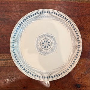 May include: A white ceramic plate with a blue floral design. The plate features a central starburst pattern and a repeating pattern of vertical lines with oval shapes. The plate is round and sits on a wooden surface.