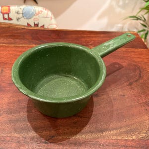 Puede incluir: Un cazo pequeño y redondo de color verde bosque con un acabado blanco moteado y un solo mango. El utensilio de cocina presenta una superficie esmaltada lisa con un patrón moteado consistente en todo el interior y el exterior, incluido el mango.