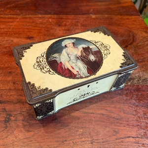 Könnte beinhalten: Eine Vintage-Blechdose mit cremefarbenem Hintergrund und goldenen Akzenten. Der Deckel zeigt ein gemaltes Bild einer Frau in einem weißen Kleid, die ein braunes Pferd reitet. Der Text auf der Dose lautet "The Millward from J. Millward & Sons Ltd."