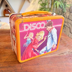 May include: Vintage metal lunchbox with an orange handle and trim. The front features a pink background with the word "DISCO" in white, and illustrations of people dancing. The lunchbox has the word "FEVER" on the top.