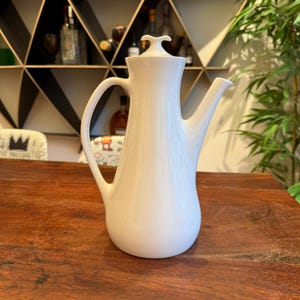 May include: A white ceramic coffee pot with a curved handle and a spout. The pot has a lid with a small handle and sits on a wooden surface. The background includes a shelf with bottles and plants.