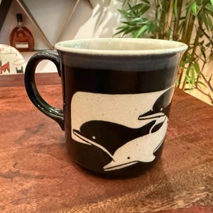 Może przedstawiać: Ceramiczny kubek z ciemnoczarnym szkliwem i kontrastującym kremowym prostokątnym panelem z grafiką przedstawiającą dwa delfiny. Kubek posiada solidne ucho oraz gładką, zaokrągloną krawędź z widoczną nakrapianą teksturą na całej powierzchni.