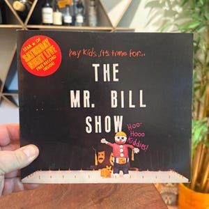 Op de afbeelding: Een zwarte platenhoes met de titel The Mr. Bill Show in witte letters. Het bevat een rode ronde sticker met de tekst Star of Saturday Night Live Free Record Inside. Een kleifiguur van Mr. Bill staat met een kleine oranje hond op een podium.