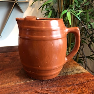 May include: A brown ceramic pitcher with a barrel-shaped body and a handle. The pitcher has a textured surface with a pattern of diagonal lines.