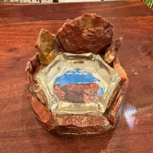 May include: A clear glass ashtray surrounded by rough, reddish-brown petrified wood pieces. The center of the ashtray features a printed image of a rock formation with the text Souvenir of Arizona and Petrified Forest, Arizona.