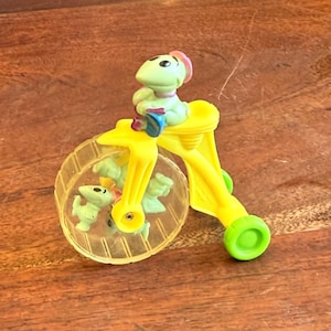 May include: A yellow toy tricycle with a green figure riding on top. The tricycle has a clear wheel with several green figures inside. The figure on top is green with a pink hat and blue accents.