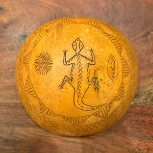 Puede incluir: Una calabaza redonda, de color marrón claro, con un diseño grabado detallado. El diseño incluye un lagarto, un sol y patrones geométricos. La calabaza tiene un aspecto y una textura naturales y orgánicos.
