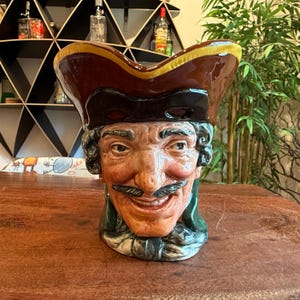 May include: A ceramic pitcher shaped like a smiling pirate's head, featuring a brown tricorn hat with a yellow trim. The face is detailed with a dark mustache and curly hair, with a green collar. The pitcher sits on a wooden surface.