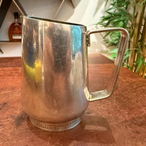 May include: A silver-toned metal pitcher with a smooth, reflective body and a rectangular handle. The base of the pitcher features a decorative band with a repeating geometric pattern. The surface shows minor scratches and signs of wear.
