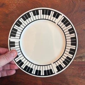 May include: A white ceramic plate with a black piano key design around the rim. The plate is round and has a smooth, glossy finish. The plate is held by a hand, and the background is a wooden surface.