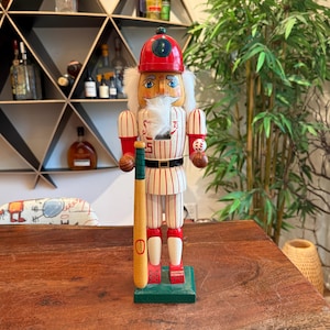 Könnte beinhalten: Ein Nussknacker-Figur im Baseball-Stil. Die Figur trägt eine Baseball-Uniform mit rot-weißen Streifen, eine rote Mütze und einen weißen Bart. Sie hält einen Baseballschläger und einen Baseball.