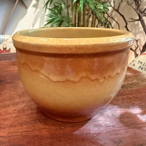 May include: A round, glazed ceramic bowl with a wide rim. The bowl has a light yellow exterior with a darker, wavy band around the middle. The interior is a lighter yellow. The bowl sits on a wooden surface.