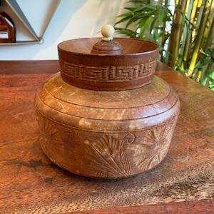 May include: A two-piece, handcrafted wooden container with a lid. The lower section is a large, round bowl with carved floral designs. The upper section is a smaller, round bowl with a geometric pattern and a small, round lid with a white knob.